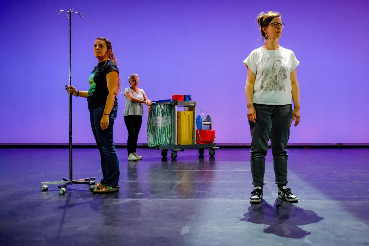 Trois interprètes immobiles sur scène, avec un pied à perfusion et un chariot de ménage, sur fond violet.