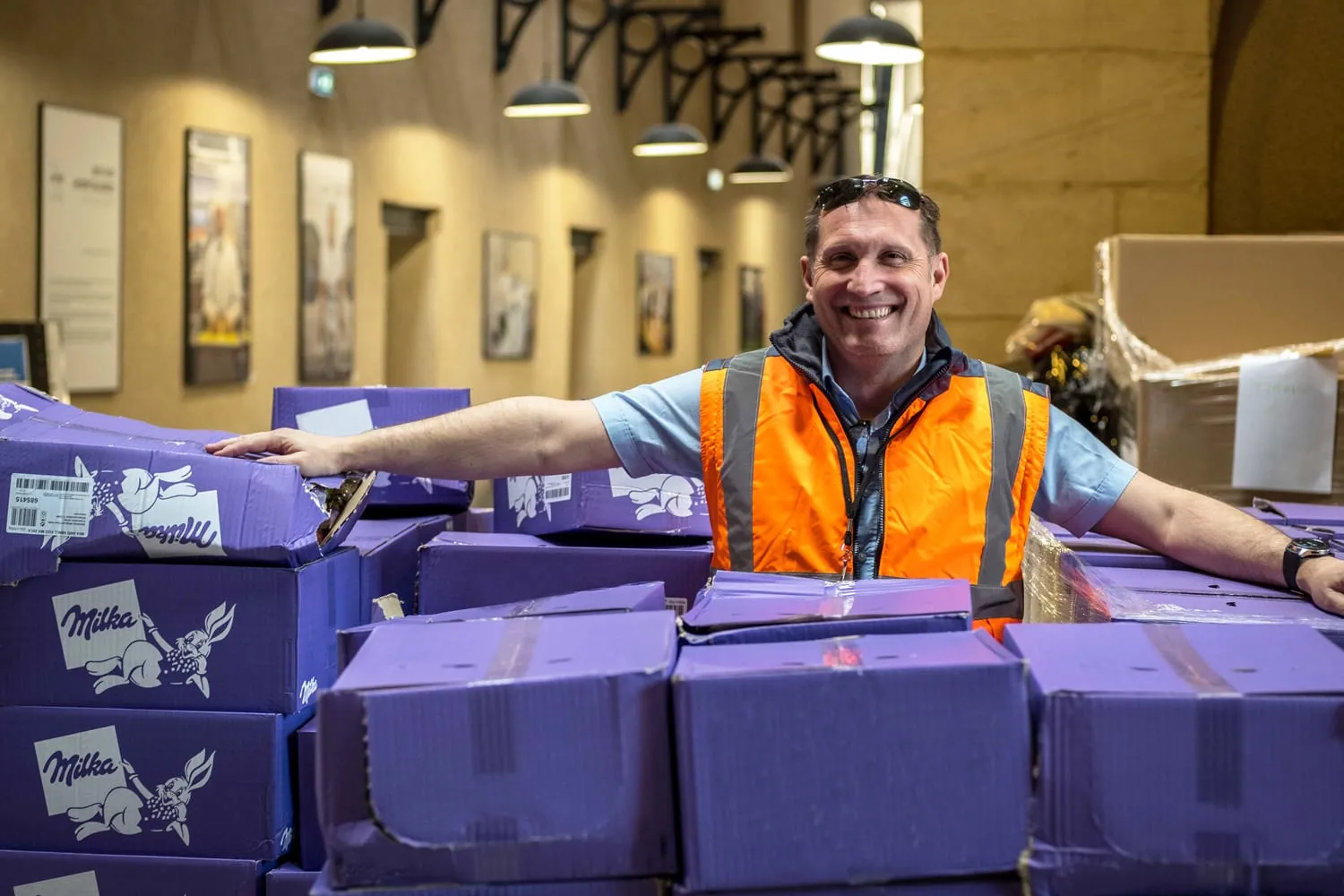 Un bénévole en gilet de sécurité derrière des palettes de cartons de chocolats destinés aux enfants hospitalisés.