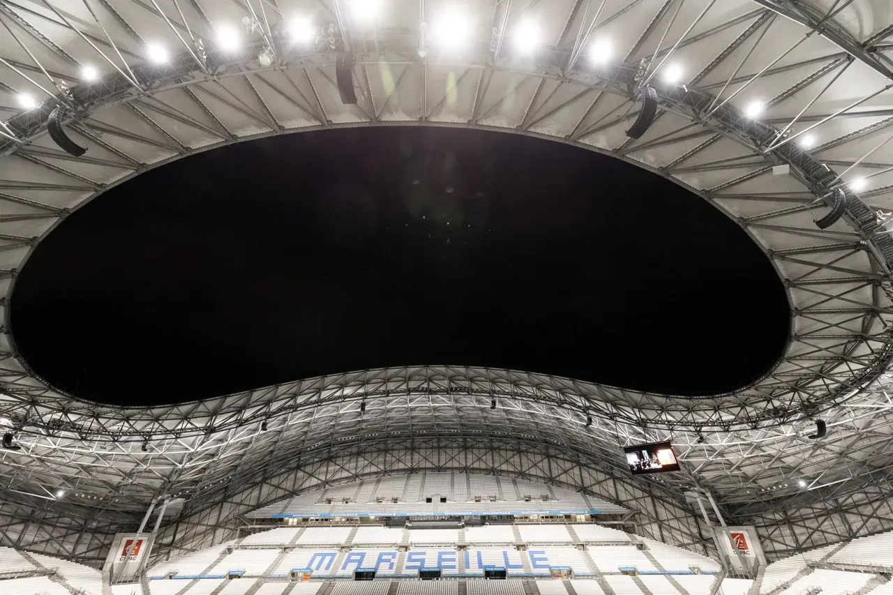 La charpente de l’Orange Vélodrome vue depuis les tribunes, le nom « Marseille » inscrit sur les sièges du virage.