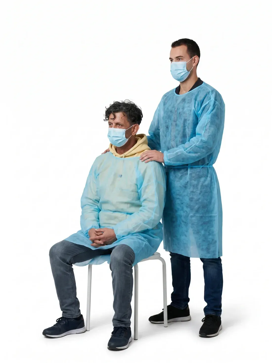 Un patient assis en surblouse et masque, la main d’un soignant debout posée sur son épaule, sur fond blanc.